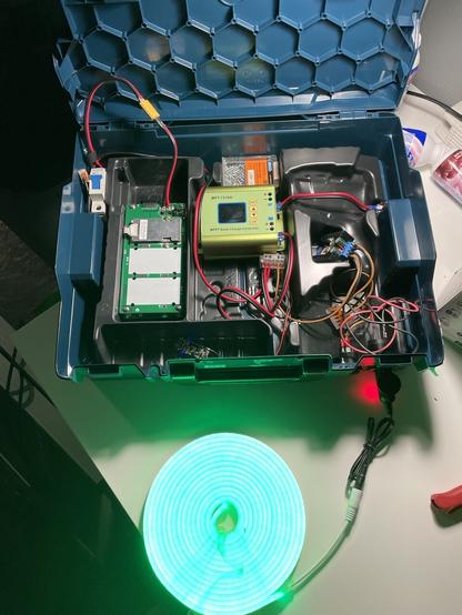 A tool suitcase containing cables and electronics. A green box, the solar charge controller, in the middle, around it a PCB covering the battery, fuses, connectors and smaller PCBs. An LED Strip is plugged into a connector on the front of the case