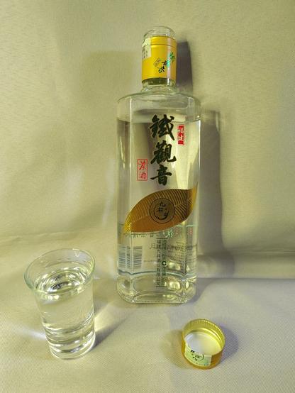 Front view of the opened bottle with a full shot glass of the contents.  From the Taobao description of the product:

九井乡福建铁观音茶酒浓香白酒正宗纯粮固态52度口粮500ml 

Jiujing Township Fujian Tieguanyin Tea Wine Luzhou-flavored Liquor Authentic Pure Grain Solid 52 Degree Ration 500ml

The speed with which the wine tears form around the walls of the shot glass are a testament to just how much alcohol is in the liquor.  This photo was shot about 15 seconds after the pour.