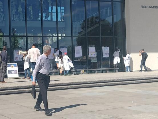 A man with a suitcase is walking while looking at scientists pasting scientific posters on the walls of Freie Universitaet in Berlin