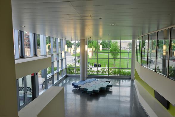 An interior view, from the mezzanine, of one of the entrances to the Integrative Learning Center, where the Journalism Department is housed at UMass Amherst. Moveable seats are arranged in an eye-catching pattern, surrounded by windows. The glass enclosure of the broadcast studio is visible on the right.