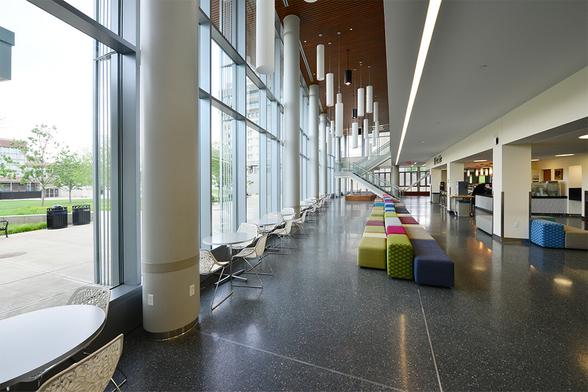 An interior view of the cafe space on the first floor of the Integrative Learning Center, where the Journalism Department is housed at UMass Amherst. Tables and fuzzy benches stand next to tall windows flanked by pillars. The coffee counter is visible in the background.