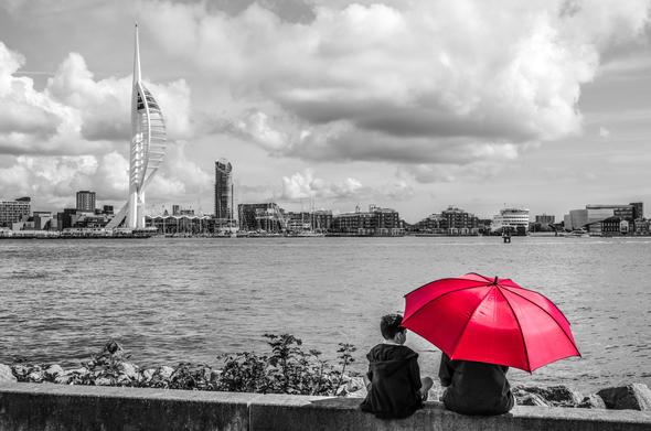 Looking over to the Spinnaker Tower from Gosport waterfront. In the background is the aforementioned tower at Gunwharf Quays. In the foreground a mother and child sitting on the harbour waterfront wall. The photo has been converted to black and white with the exception of a red umbrella being used as a sunshade by the mother.