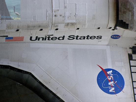 A photograph looking down over the port side wing of NASA’s Space Shuttle. The top of the image shows the port side of the fuselage with the US flag and the words UNITED STATES printed on the side with a blue NASA circular logo. The wing stretches down the bottom of the image, showing its white upper surface and the larger blue NASA “meatball” logo painted on prominently.