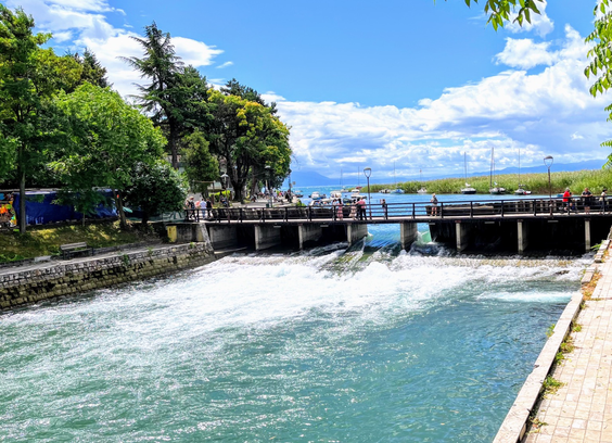 Water flow under a bridge from a lake behind. Greenery on all sides of water. Tourists on the bridge. Blue skies filled with White clouds above.