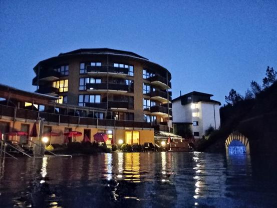 Foto aus Pool Richtung Hotelgebäude zu blauen Stunde
im Vordergrund spiegel sich die Lichter der Terrasse im Wasser, im Hintergrund ein beleuchtetes rundes 5 stöckiges Hotel