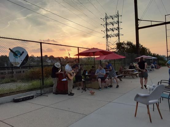 Musicians jam by outdoor tables on path to bridge. Guitar, ukulele, clarinet and more.