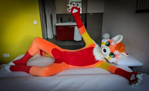 Skin-tight red panda fursuit with a red/orange/yellow/white color scheme, with green anime-style eyes. He's lying on a bed, rolled over to face the camera, both arms extended out to welcome hugs and snugs.