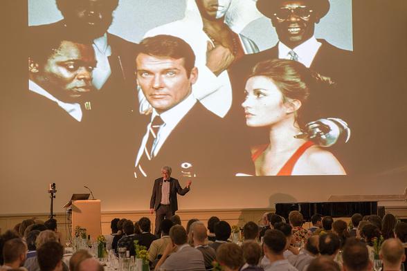Metin Tolan vor einer Folie mit James Bond + weiteren Figuren.