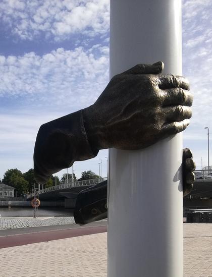 Lähikuva edellisessä kuvassa näkyneistä käsistä, jotka pitelevät kiinni valkoisesta lyhtytolpasta.