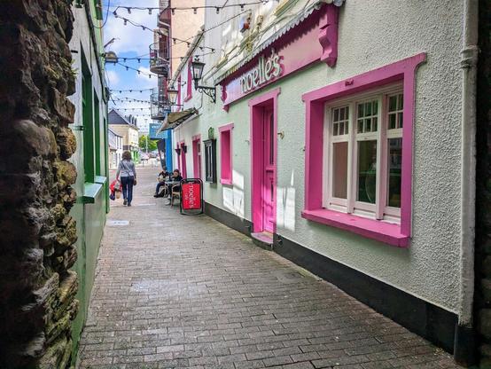 On the left, there is a stone wall, followed by a green storefront.  On the right, there is a white storefront with pink accents around the windows, a pink door, and a pink sign that reads "Noelle's".  There is a footpath down the middle of the image.  In the distance, you can see people drinking coffee and hanging out at a series of outdoor tables.
