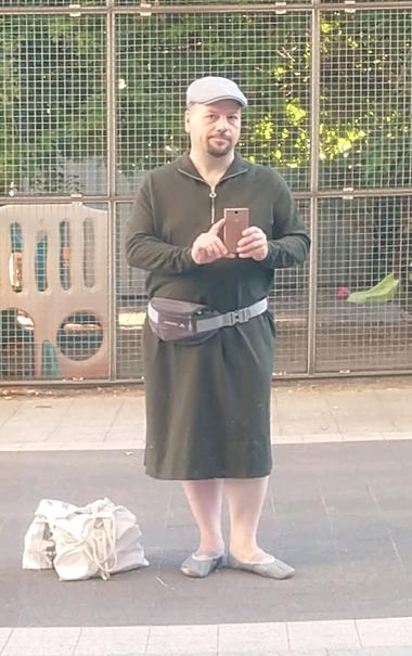 Selbstporträt in verspiegeltem Fenster in einer Fußgängerzone. Mann mittleren Alters mit dunklem Kinnbart in wadenlangem olivgrünen Kleid. Schlichter gerader Schnitt, lange Ärmel, oben mittig ein halboffener Reißverschluss, Stehkragen mit umgeklappten Ecken. Dazu in hellem grau eine Bauchtasche, Schirmmütze und Ballerinas mit Kreuzgummi.