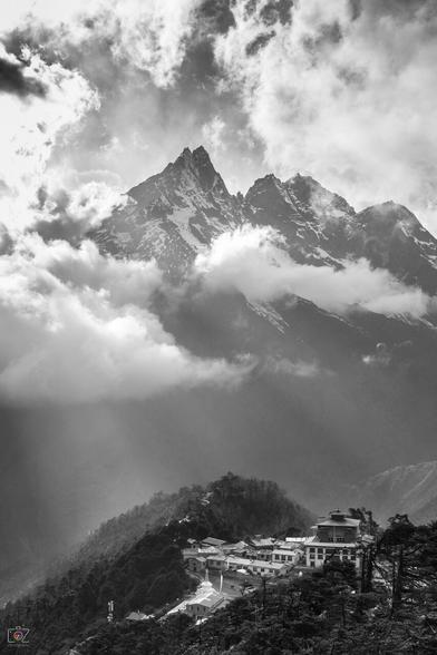 A black and white photo. On the lower right you can see some buildings of a Buddhist monastery on a hill, surrounded by trees. Behind it, high mountains with rugged peaks rise through the clouds. Some decent rays of sunlight fall on the monastery through these clouds.