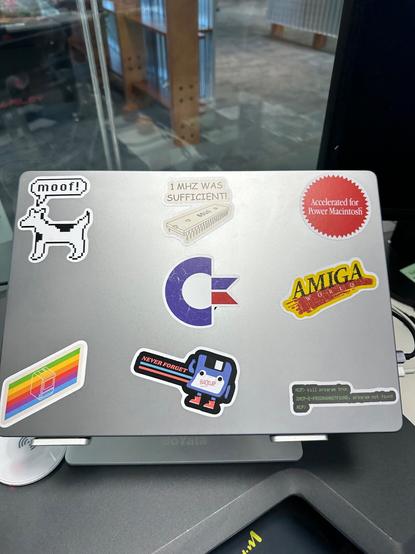 Picture of the top of my laptop with retro computing inspired stickers like a commodore logo, amiga world, Clarence the dogcow etc.