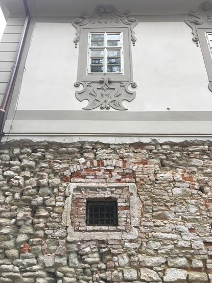 The first floor of this building is a jumble of roughly textured rocks of different sizes forming the wall, which seems to have been made in the 18th, maybe even 17th century. Set into the wall is a small square window, blocked by a grid of iron bars, and framed by bricks and (mortar?) that appears to have been set into the wall at a later date - perhaps mid to late 18th century? The second story is radically different, perhaps late 19th Century; a smooth, elegant gray wall, with offsetting gray trim.set in the middle of the wall is a glass paned window, framed by an elaborate gray, abstract scroll design.