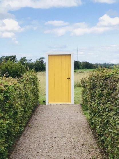 A framed yellow door sits at the end of a path between two rows of hedges. Behind the door is an open meadow.