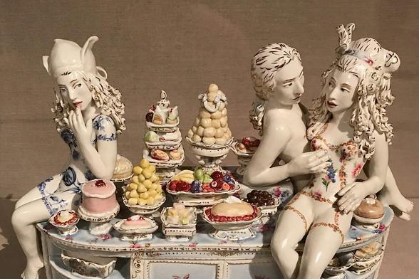 A porcelain sculpture titled 'Sideboard' by Chris Antemann, featuring an intimate couple perched sensuously on a sideboard, surrounded by tempting desserts and fruits. The female figure gazes provocatively at another woman on the opposite side, exuding an air of intrigue and desire.