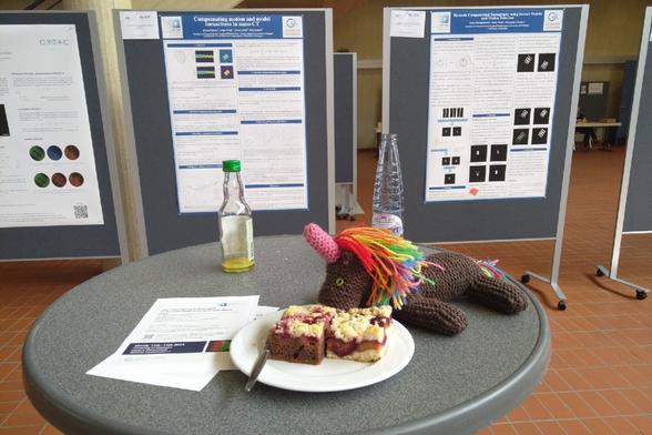 1 Strickhorn mit 2 Kuchenstückchen auf Stehtisch vor Posterstellwänden.