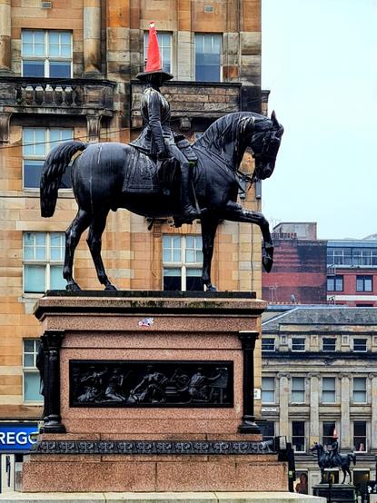 Two statues of a man on horseback, one in the foreground and one in the bakcground, both with road cones balanced on their heads.