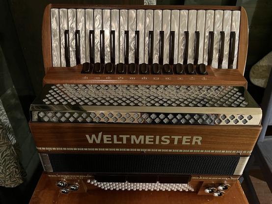 Photo shows a accordion made of light brown wood, branded “Weltmeister” with white and black pearlized keys
