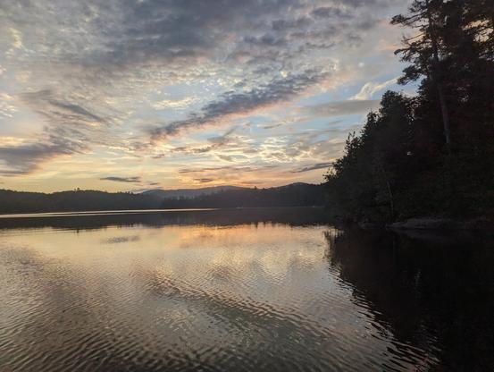 Lake reflecting trees, mountains in distance, and clouds lit orange by sunrise