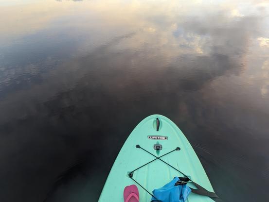 Front of standup paddleboard on lake reflecting sky and clouds