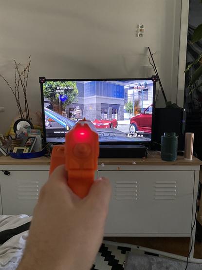 Ma main tenant le pistolet orange « Gun-Con 3 » devant la télé affichant le jeu « Time Crisis 4 » sur PS3.