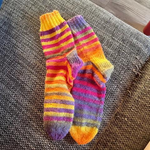 A pair of hand knitted socks with horizontal stripes. The base color is a bright yet warm yellow. The stripes are in different shades of violet and pink.