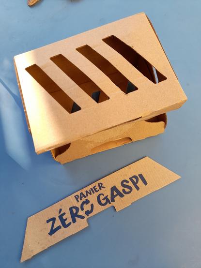 A cardboard fruit box with openings. A cardboard strip with the French inscription "Panier zero gaspi" = Zero waste basket.