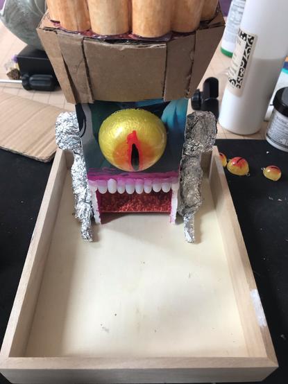 photograph of a dice tower in a dice box. The tower has tin-foil armatures on the sides, a large epoxy resin eye in the center, and teeth where the dice will exit the tower.
