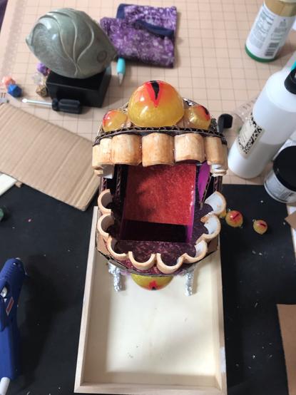 Photograph of the top of a dice tower. Three eyes crown the opening, on top of a set of blunt teeth where the dice go in.