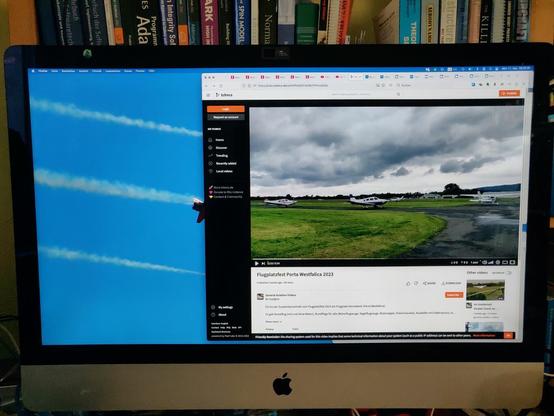 Photo of a 27" iMac running Firefox showing a peertube video of light aircraft lining up for takeoff. A book shelf in the background with various programming and aviation-related titles.