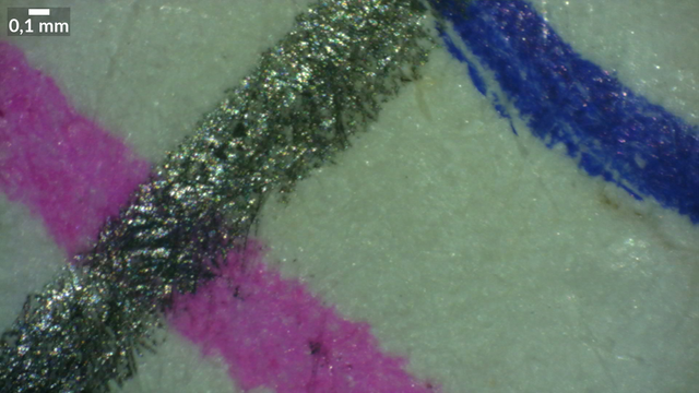 Mikroskopaufnahme 3,4mm x 1,9mm. Die Faserstruktur eines weißen Papiers ist zu erkennen, darauf rechtrs oben ein Strich mit Kugelschreiber in blau, links unten ein Strich mit einem Stabilo Point 88 in rosa und quer darüber ein Strich mit einem Bleistift.