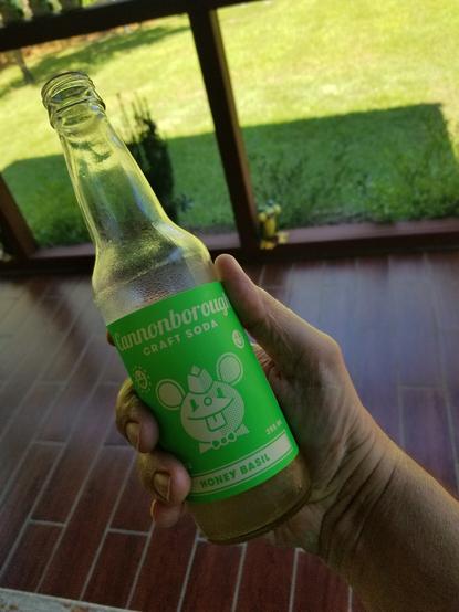 Consumer holding the last few ounces of Cannonborough Honey Basil soda.
(Unobtanium Edition)
Product shown in a clear glass soda bottle (12oz).  The label has a green background with white for logo and text.
Company name "Cannonborough" is written in cursive.  The mascot of the Honey Basil blend is a short-eared rodent with a soda jerk uniform, depicted in a headshot.
Beneath the label, the product is visible as a sort of light tea colored translucent liquid.