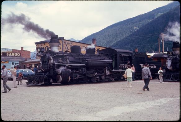 A steam locomotive - black with silver and white accents - stands at the end of the track in the middle of a dirt road (facing left). Smoke drifts to the left from its diamond-shaped smokestack and wisps of steam are visible near the cylinders and at its top. A few tourists stand around the locomotive, observing and taking pictures. Beyond stands a yellow frame building with painted signs reading “Old Arcade.” In the distance, a tree-covered mountain rises above the town.