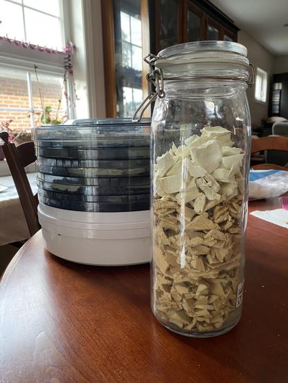 Big jar of crumbled, dried puffball in front of a dehydrator full of mushroom slices. The pieces at the top of the jar are white, but the pieces at the bottom are tan.