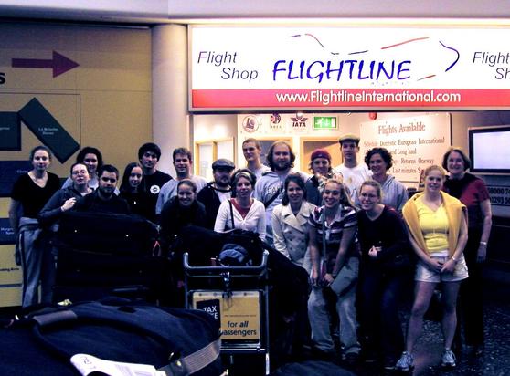 A group of 19 college students and one professor who have just flown overnight from the United States stand together amongst luggage carts and other airport trappings in London Gatwick. The photo is poorly exposed due to backlighting from a large sign that reads “Flightline Flight Shop www.FlightlineInternational.com”