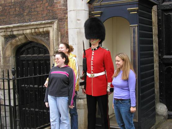 Three young women stand around a Grenedier Guard outside of St James’s Palace. The guard, in traditional red coat and black bearskin, stands at attention. The three women look towards another photographer (out of frame). The tallest of the three women, partly obscured by another of them, wears a yellow windbreaker a with white stripe down the arm.