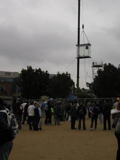 A small crowd is gathered, looking up at a plexiglass box that hangs from a crane. Inside the box, a man reclines, partially covered by a sheet.