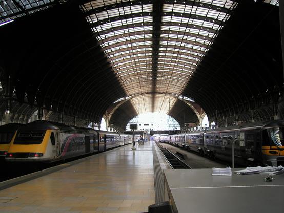 View down platforms 4 & 5 of Paddington Station. A steel and glass roof arches overhead, and trains sit waiting at each platform.