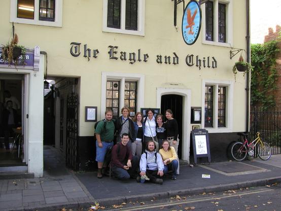 A group of nine college students pose for a photo in front of the Eagle and Child pub. The building is painted pale yellow, and the fancy black letters spelling out “The Eagle and Child” are affixed to the façade. A painted sign depicting an eagle in flight carrying a baby in a sling held in its claws hangs from the front of the building.