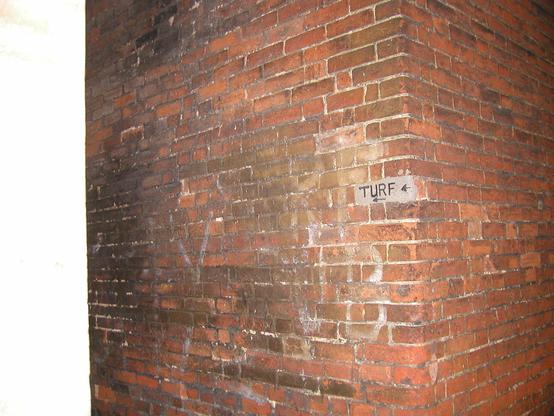 Two brick walls meet at a corner, the point of which is towards the camera. The space of a single brick has been mortared over, and on that gray surface is written the word “TURF” and two arrows that point the way into the alley and towards the pub in question.