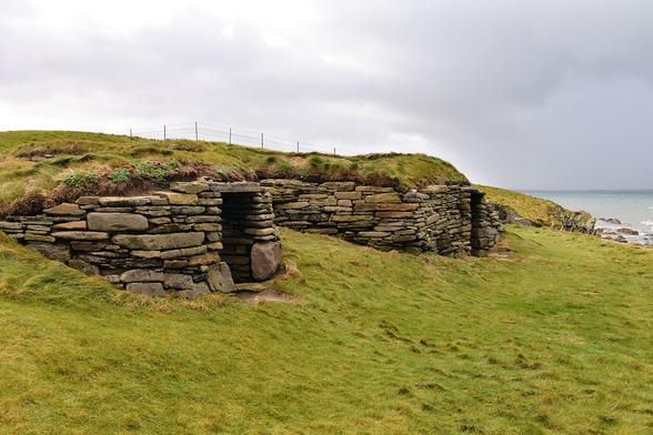 Two low stone walls with doorways set in a grassy covered slope beside the sea. Very overcast.
