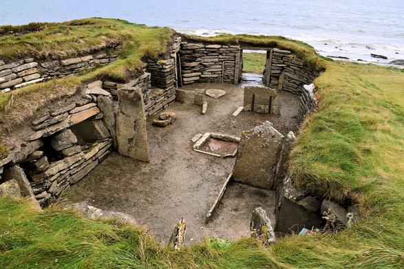 From a grassy bank looking down into a a stone-walled room with dirt flower. Upright flat stones divide the space. The nearest space has squares set into the stone wall (cupboards). The middle section has a square in the floor edged by stone (hearth). In the furthest wall, a low doorway leads outside, towards the sea.