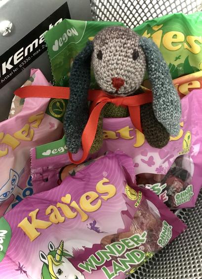 Kleiner Häkel-Hase mit roten Näschen, roter Schleife um den Hals und freundlichem Gesichtsausdruck sitzt zwischen fünf Tüten Fruchtgummis in einem Fahrrad-Einkaufskorb und hält die Tüten mit Pfötchen und Ohren fest.