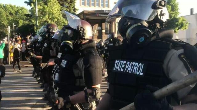 A photograph of armoured riot cops in a line, wearing gas masks. Their uniforms read STATE PATROL.