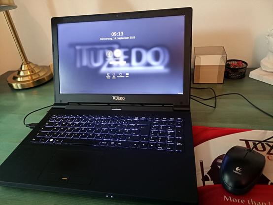 Aufgeklappter Laptop mit Linux-Betriebssystem "Tuxedo OS" auf grünem Schreibtisch.