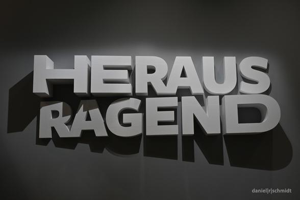 Auf einer anthrazitfarbenen Museumswand stehen weiße Reliefbuchstaben heraus. Sie sind mit einem Spot in Szene gesetzt und ergeben das Wort "Herausragend".