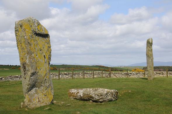 Drumtroddan Standing Stones. The image shows a rough grassy field with a stone wall in the background and distant views beyond that. On the left of the image is a large standing stone covered in lichen and there is another standing stone, a little more distant, on the right of the frame. A third stone is lying on the ground in the bottom middle of the frame. The scene is in sunlight.
