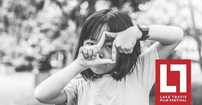 A B&W photo of a girl holding up her hands looking through her fingers in a vertical aspect of a film screen.