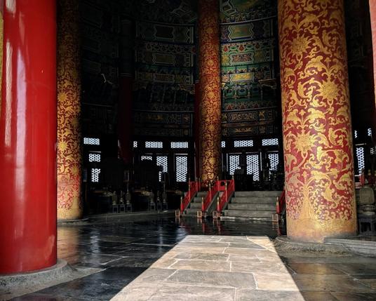 This is the interior of a large, wooden structure.  All light sources appear to be natural and light is pouring from the doorway.  Tall red pillars are visible, coming out of a stone floor.